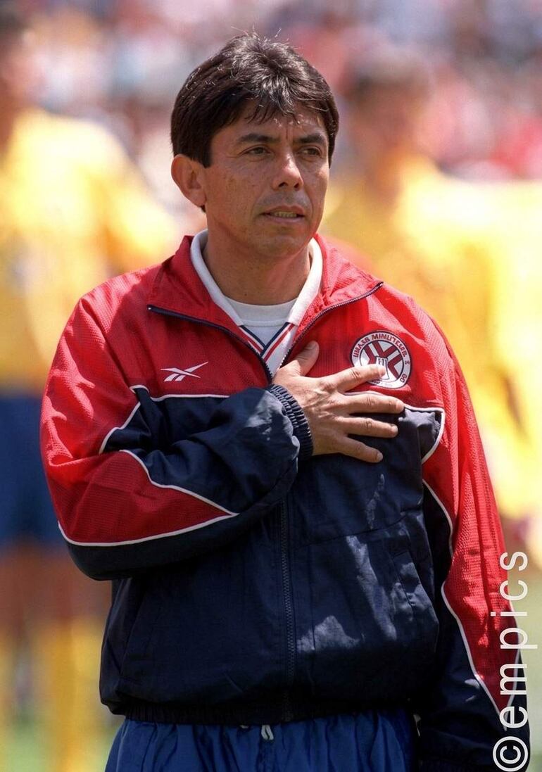 Carlos Morales Santos con chaqueta roja y azul, mano sobre el corazón en un entorno deportivo.