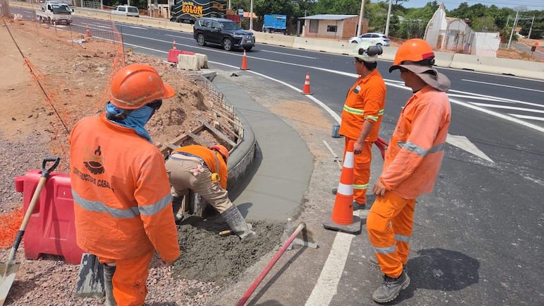 Trabajador agachado aplicando cemento, rodeado de tres colegas en camisetas naranjas, en obra vial con maquinaria de fondo.