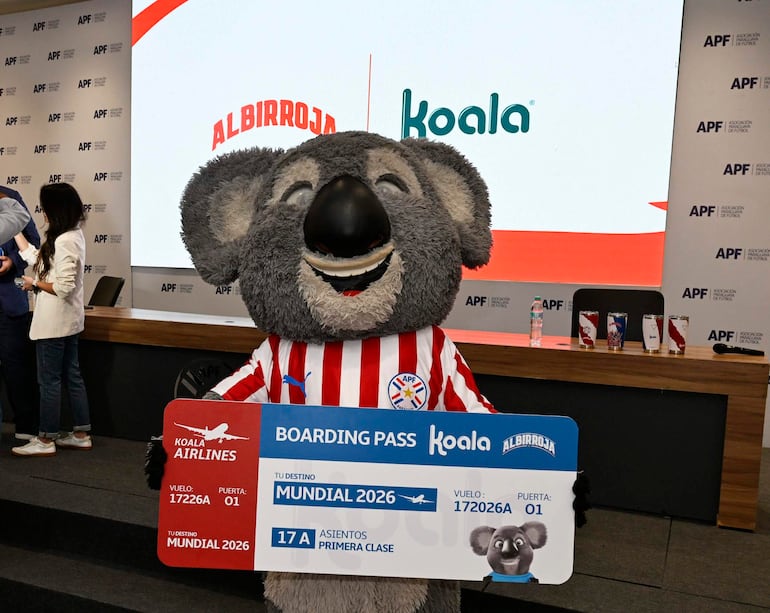 Figura en disfraz de koala sostiene un pase de abordar con destino Mundial 2026 en un salón con dos personas al fondo.