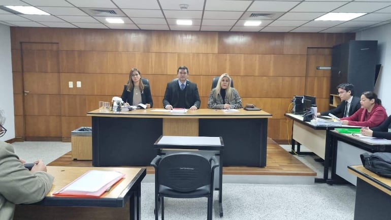 Darío Javier Báez en mesa de tribunal con Natalia Cacavelos y Gloria Hermosa, todos observando documentos en una sala formal.