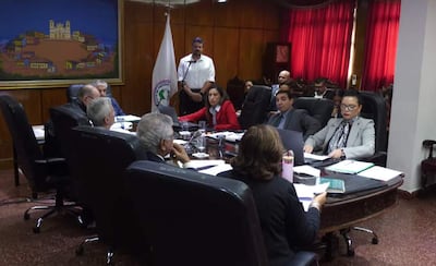 Bettina Albertini en abrigo rojo y Juan Carlos Frutos observan atentamente en reunión formal con gente alrededor y decoración institucional.