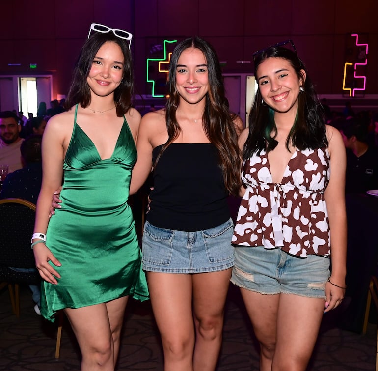 Tres jóvenes posan sonrientes: una con vestido verde, otra con camiseta negra y pantalones cortos, y otra en blusa marrón y shorts, en un ambiente festivo.