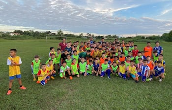 Cien niños organizados en fila en un campo de fútbol, bajo cielo parcialmente nublado, mientras un adulto supervisa la actividad.