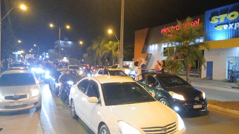 Dos personas celebran sobre un vehículo en una multitudinaria caravana nocturna. Los coches tienen las luces encendidas.