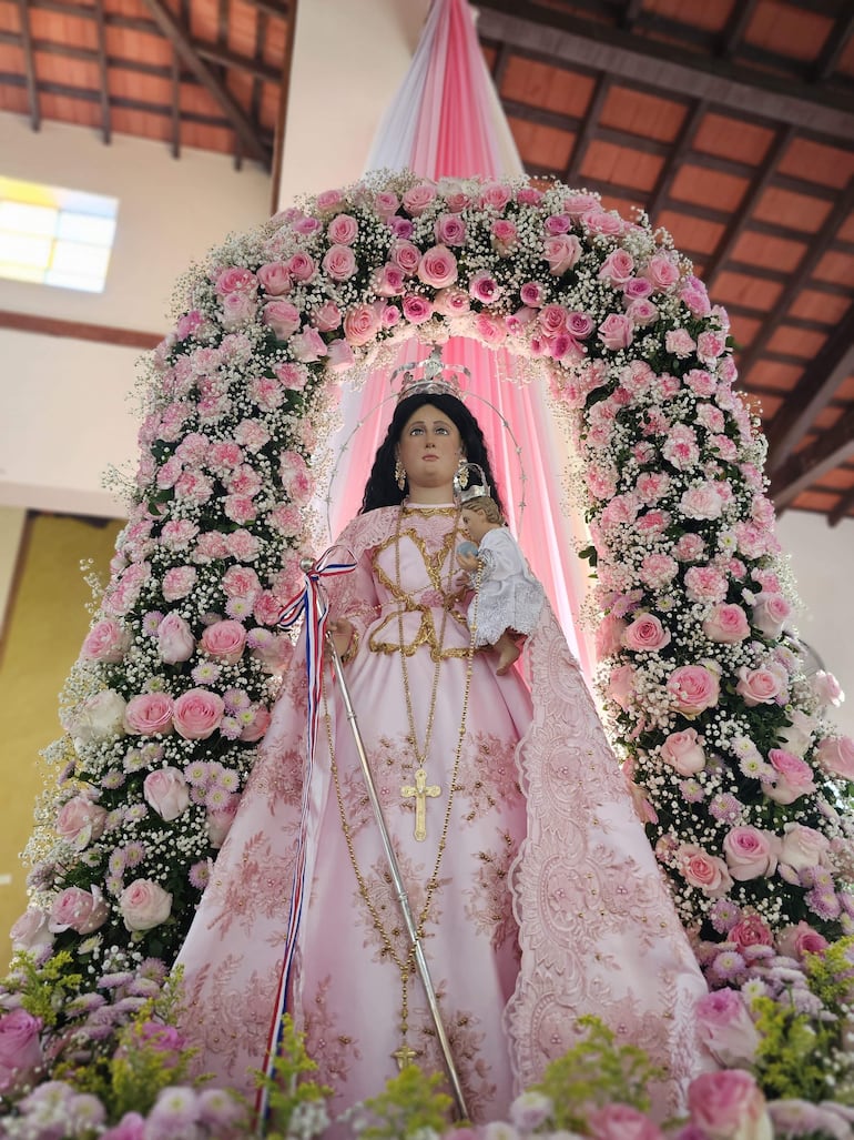 Virgen del Rosario vestida con túnica rosa y corona, rodeada de flores rosadas en un ambiente solemne.