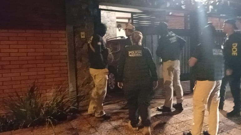 Agentes de la Policía Nacional en la entrada de una vivienda, vestidos de negro, conversando y en postura de vigilancia en operativo nocturno.