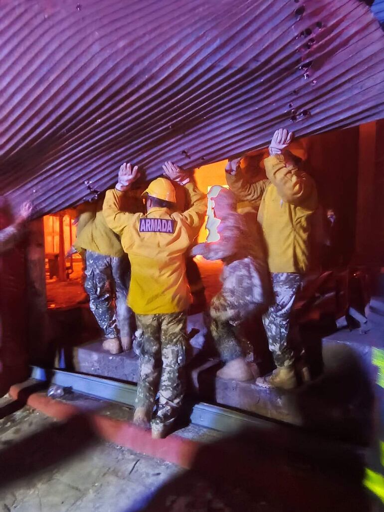 Seis personas vestidas con chaquetas amarillas y pantalones de camuflaje levantan una estructura metálica en un entorno oscuro y tenso.