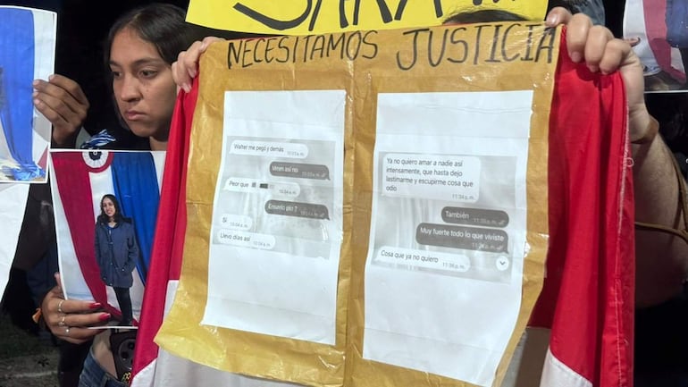 Mujer joven con cabello oscuro sostiene cartel llamativo 'Necesitamos justicia' en protesta, rodeada de fotos y mensajes de denuncia.