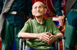 Hombre mayor en silla de ruedas, camiseta verde y pantalones azules, flanqueado por jóvenes soldados en uniformes verdes, decoraciones con globos rojos y dorados.