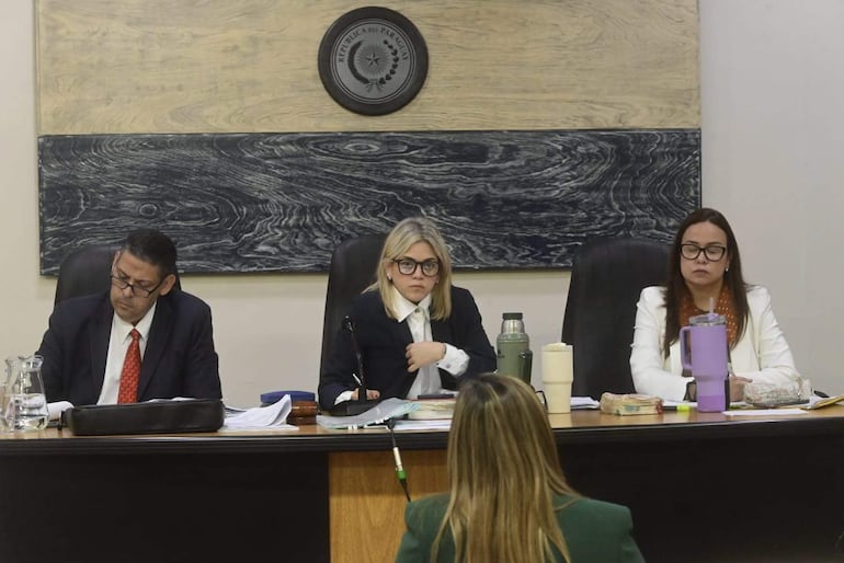 La mujer de gafas y vestido oscuro escucha atenta mientras dos acompañantes observan en el juzgado decorado con el escudo nacional.