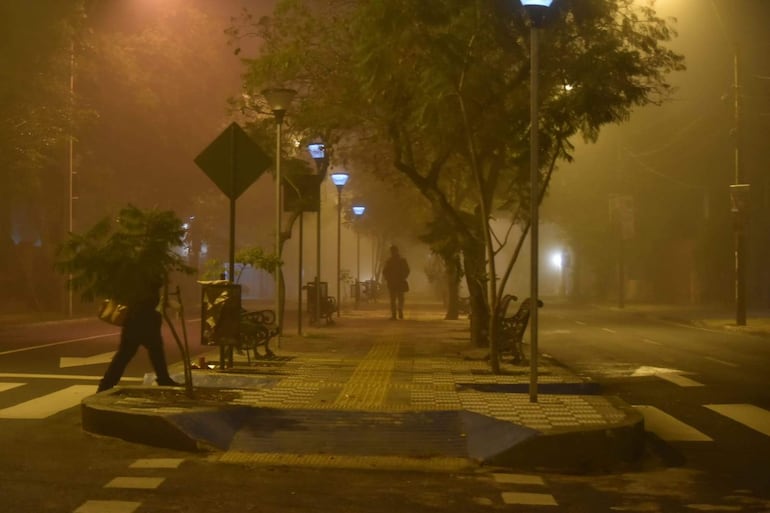 Dos personas vestidas casualmente caminan en una calle brumosa, iluminada por lámparas tenues en una atmósfera tranquila.