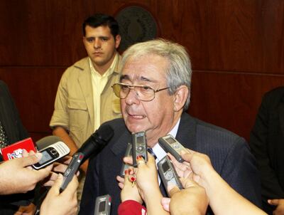 Hombre mayor con gafas habla a periodistas. Varias manos sostienen micrófonos. Ambiente de atención mediática.