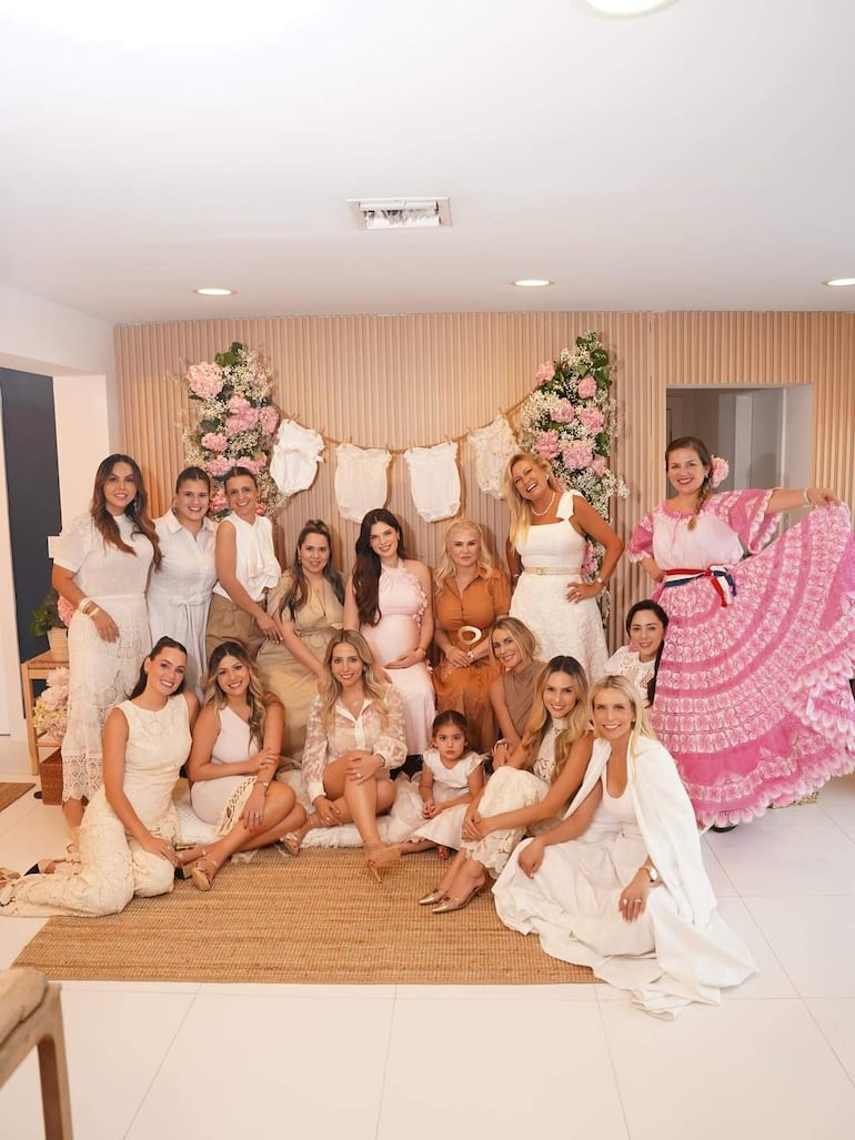 Grupo de mujeres y una niña sonrientes, vestidas con tonos claros y un vestido rosa tradicional, en un ambiente decorativo festivo.