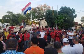 Grupo de personas reunidas en el exterior, con un hombre de traje oscuro dirigiendo la conversación, mientras ondea una bandera paraguaya.
