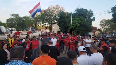 Grupo de personas reunidas en el exterior, con un hombre de traje oscuro dirigiendo la conversación, mientras ondea una bandera paraguaya.