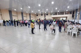 Asistentes con gorras verdes en fila, algunos revisan documentos y otros usan teléfonos móviles en un ambiente colaborativo.