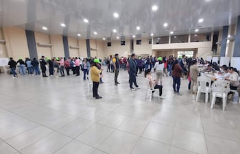 Asistentes con gorras verdes en fila, algunos revisan documentos y otros usan teléfonos móviles en un ambiente colaborativo.
