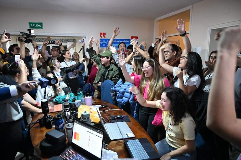 Jóvenes emocionados en el PC de Patria Querida, una mujer sonriente con camiseta clara y un hombre en chaqueta verde habla alentador.