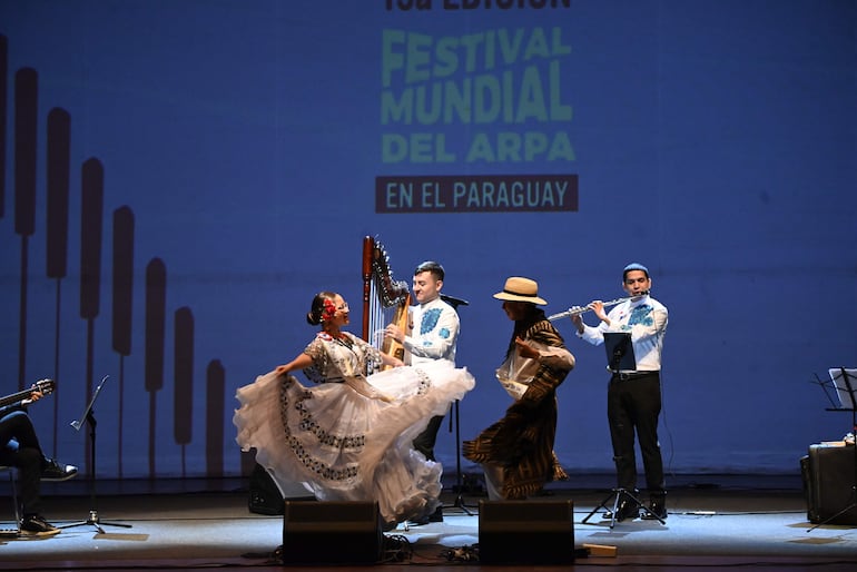 Mujer en traje tradicional baila en el escenario, mientras un hombre toca el arpa y otro la flauta, ambiente festivo.