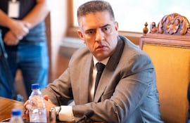 Hombre en traje gris oscuro y corbata blanca sentado en mesa de reunión, mirando atentamente a su interlocutor.