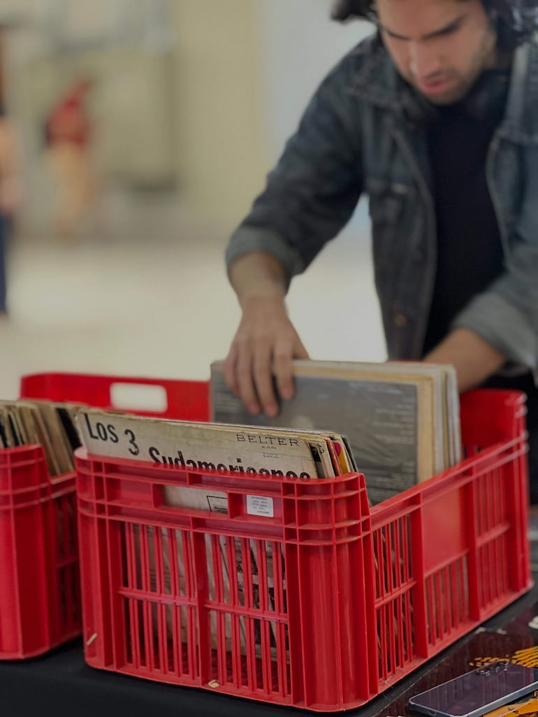 Joven con abrigo oscuro y auriculares revisa discos de vinilo en feria cultural con ambiente interior.
