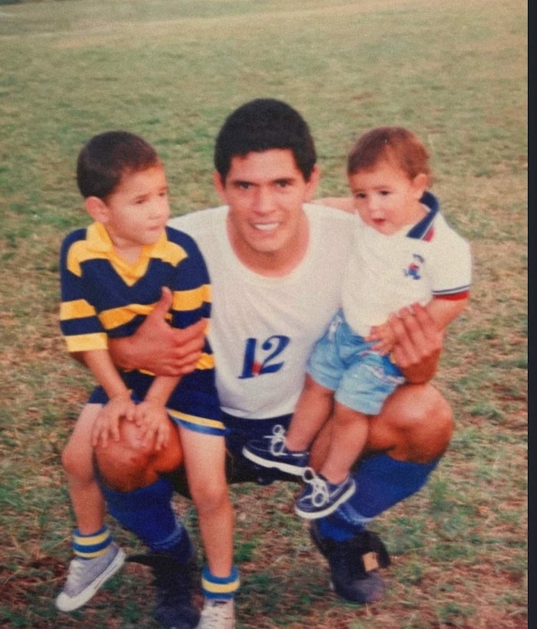 Jugador con camiseta blanca y número 12 sostiene a dos niños, uno en camiseta a rayas amarillas y azules, en un campo de césped.