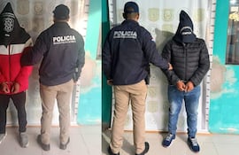 Dos hombres esposados, uno con chaqueta roja y capucha negra, otro con abrigo negro, en un ambiente policial con un oficial de perfil.