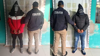 Dos hombres esposados, uno con chaqueta roja y capucha negra, otro con abrigo negro, en un ambiente policial con un oficial de perfil.