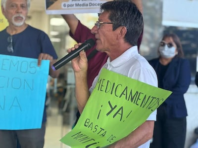 Hombre con micrófono y cartel verde 'MEDICAMENTOS ¡YA!' se dirige a un grupo de manifestantes en el Hospital.