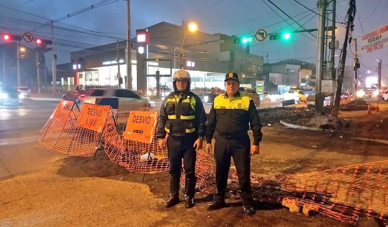 Dos agentes de tránsito en uniforme negro y amarillo regulan el tráfico, con vehículos y señalización de desvío visibles en el fondo.