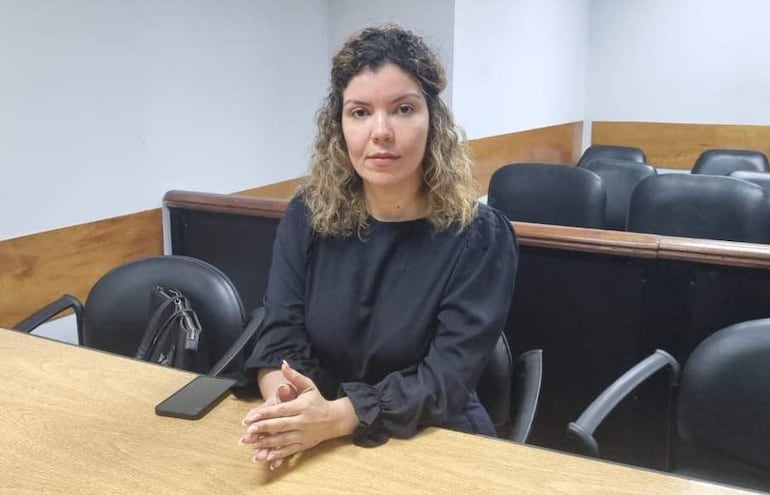 Mujer con cabello rizado y blusa oscura, sentada en una mesa de sala de reuniones, en postura reflexiva.