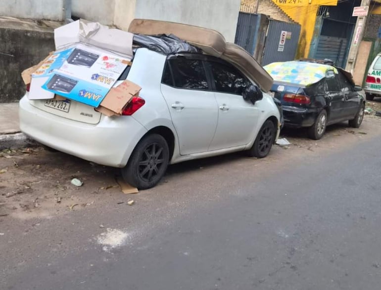 Automóvil blanco y negro cubiertos con cartones y plásticos, estacionados en un entorno urbano con hojas caídas alrededor.