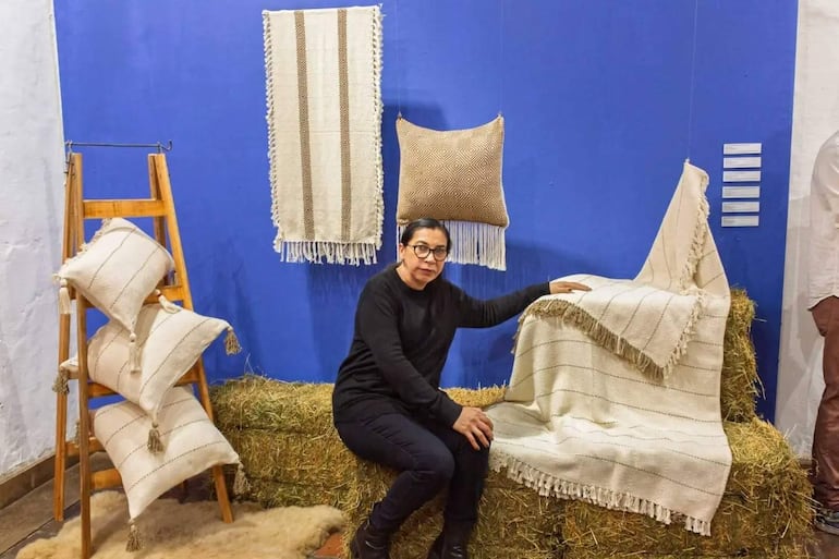 Mujer con cabello recogido y gafas, vestida de negro, sentada en un sofá con textiles coloridos a su alrededor.