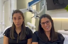 Dos mujeres con camisetas negras, una sonriente y la otra seria, conversan en un entorno profesional con elementos médicos detrás.