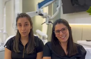Dos mujeres con camisetas negras, una sonriente y la otra seria, conversan en un entorno profesional con elementos médicos detrás.