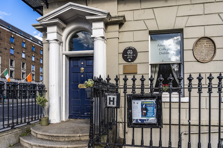 Fachada del American College Dublin con puerta azul, columnas blancas, jardín decorativo y placas que honran a Wilde.