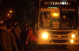 Grupo de personas de pie y entrando a un autobús iluminado, algunas con ropa casual y una gorra blanca entre ellos.