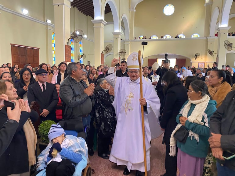 Clérigo en vestimentas litúrgicas blancas interactúa con fieles, algunos en oración, mientras un niño en silla de ruedas observa.