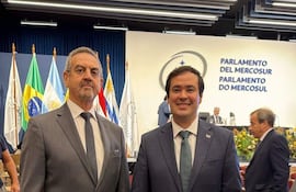 Yamil Esgaib y Rodrigo Gamarra en trajes oscuros, sonriendo en un evento formal con banderas de varios países en el fondo.