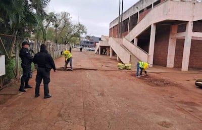 Agentes de seguridad en uniforme oscuro observan mientras hombres con chalecos amarillos limpian el suelo en mal estado.