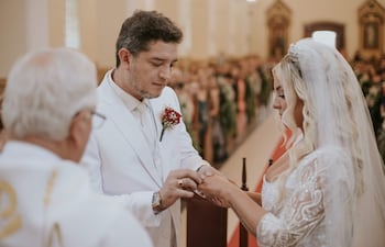 Hombre en traje blanco coloca anillo a mujer de perfil con vestido de novia, mientras sacerdote oficia ceremonia en interior decorado.