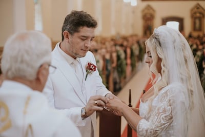 Hombre en traje blanco coloca anillo a mujer de perfil con vestido de novia, mientras sacerdote oficia ceremonia en interior decorado.