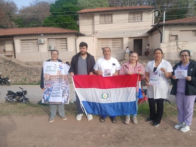 Grupo de médicos sostiene pancartas y bandera de Paraguay, posando frente a edificio relacionado con salud.