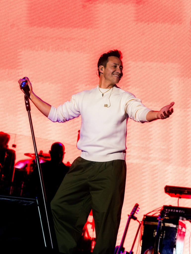 Luciano Pereyra sonríe mientras canta en un escenario iluminado, vestido con suéter blanco y pantalones oscuros, interactuando con el público.