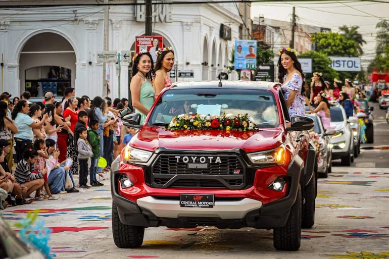 Tres mujeres sonrientes sobre una camioneta roja decorada con flores, rodeadas de una multitud animada en un entorno festivo.