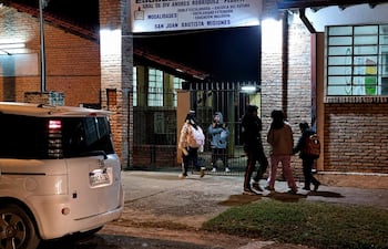 Cinco padres se reúnen cerca de la escuela con expresión de preocupación, observando las instalaciones iluminadas por la noche.