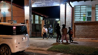 Cinco padres se reúnen cerca de la escuela con expresión de preocupación, observando las instalaciones iluminadas por la noche.
