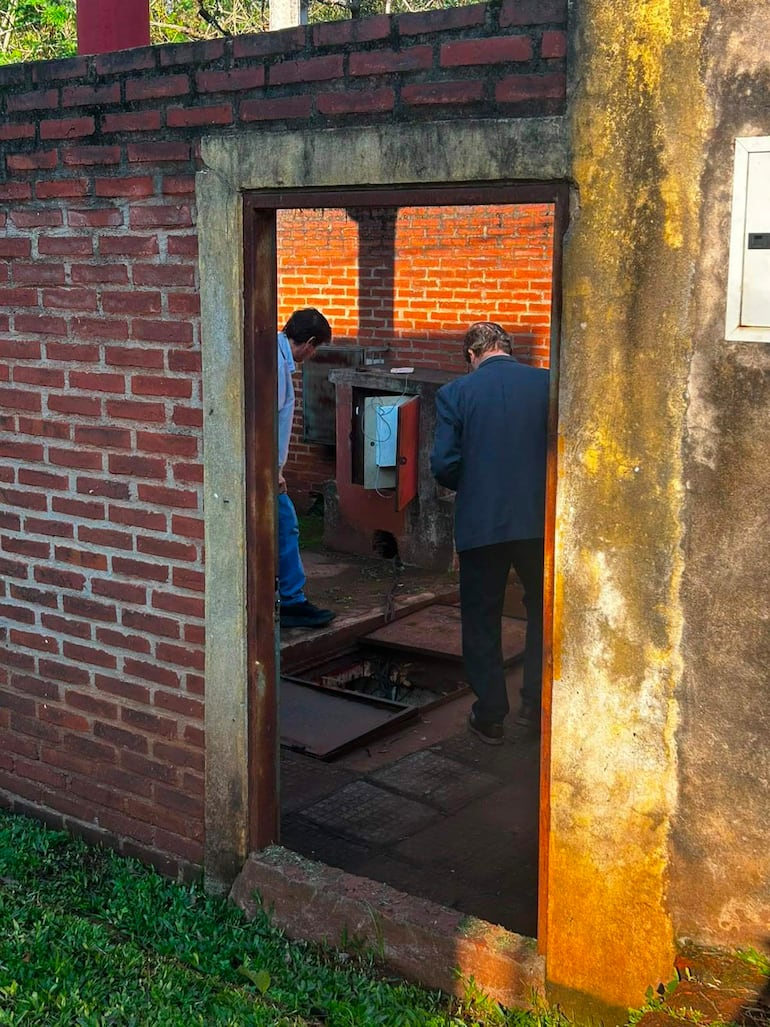 Dos hombres, uno con traje oscuro y otro en camisa blanca, inspeccionan una tapa de registro en un entorno rural.