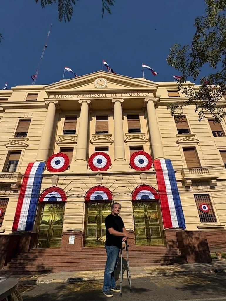 Hombre de pie con muletas, frente al Banco Nacional de Fomento, rodeado de banderas de Paraguay en un día soleado.