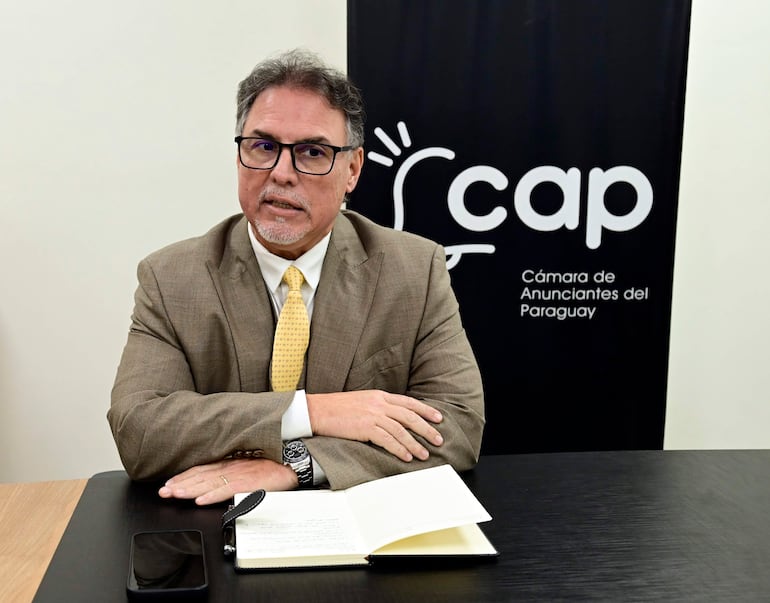 Hombre de cabello canoso y gafas, vestido con traje claro y corbata amarilla, sentado en mesa con cuaderno y teléfono apagado.